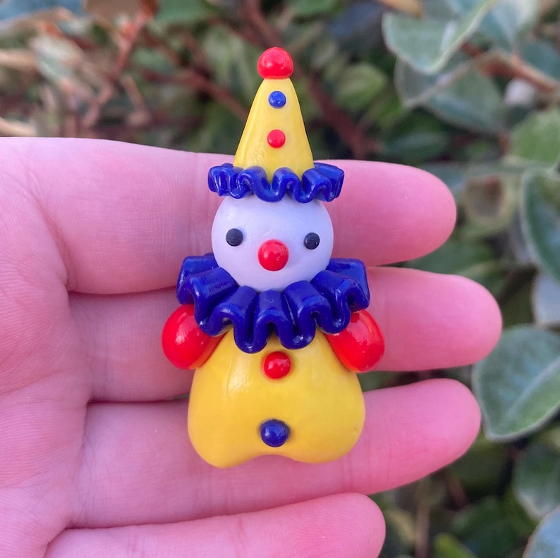 May include: A small, colorful clown figurine with a yellow body, a white face, and a pointed yellow hat. The clown has a red nose, red hands, and a blue ruffled collar. The hat is adorned with red and blue dots.