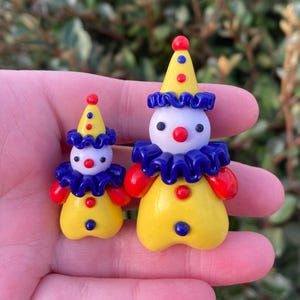 May include: Two small, colorful clown figurines. Each clown has a yellow body, red hands, and a yellow hat with blue and red accents. They have white faces with red noses and blue ruffled collars. The clowns are approximately 2 inches tall.