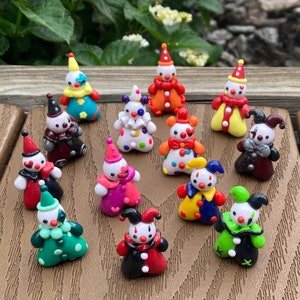 May include: A collection of small, colorful clown figurines. Each figurine has a unique design, with different colored hats, outfits, and facial expressions. The clowns are arranged on a brown wooden surface, with green foliage in the background.