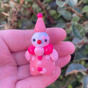 May include: A small, handmade clown figurine. The clown is primarily pink with white polka dots and a pink conical hat. It has a light blue face with black eyes, a red nose, and a pink heart-shaped detail. The clown has a pink ruffled collar.