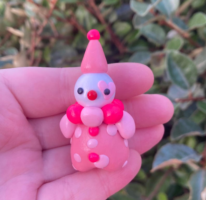 May include: A small, handmade clown figurine. The clown is primarily pink with white polka dots and a pink conical hat. It has a light blue face with black eyes, a red nose, and a pink heart-shaped detail. The clown has a pink ruffled collar.