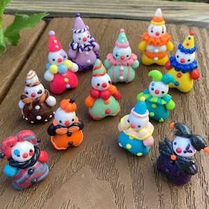 May include: A collection of colorful, handcrafted clown figurines. Each figurine features a white face, a pointed hat, and a unique outfit with vibrant colors like red, blue, and green. The clowns are arranged on a wooden surface.
