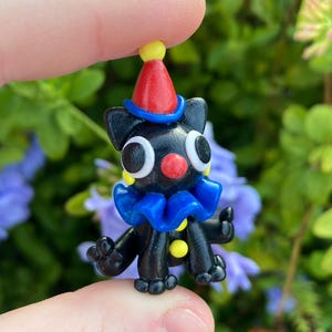 May include: A small, black figurine of a clown-like creature with large white eyes, a red nose, and a blue ruffled collar. It wears a red and yellow pointed hat. The figure is held between fingers against a backdrop of green foliage and purple flowers.