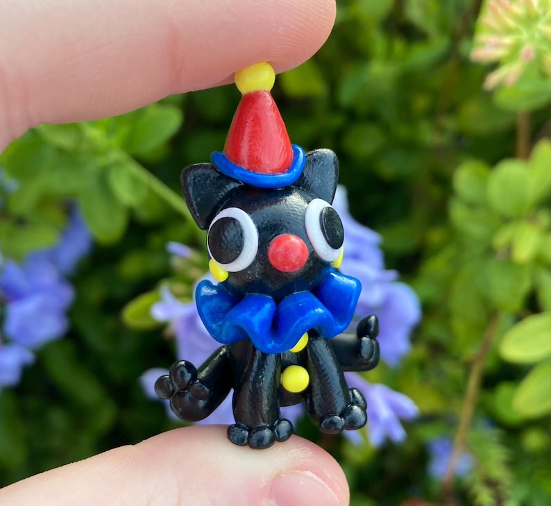 May include: A small, black figurine of a clown-like creature with large white eyes, a red nose, and a blue ruffled collar. It wears a red and yellow pointed hat. The figure is held between fingers against a backdrop of green foliage and purple flowers.
