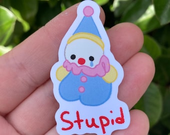 May include: A sticker featuring a cartoon clown with a blue outfit, a pink ruffled collar, and a red nose. The clown has a sad expression with a single tear. The word "Stupid" is written in red at the bottom of the sticker.
