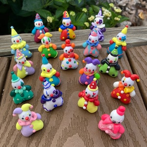 Peut inclure: Plusieurs figurines de clowns miniatures colorés. Chaque figurine présente un visage blanc, un chapeau pointu et un col à volants. Les clowns sont vêtus de différentes couleurs, dont le rouge, le jaune, le bleu, le vert et le violet, avec des pois.