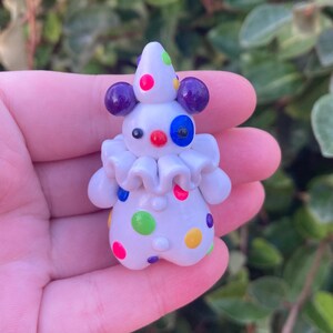 May include: A white clown figurine with a cone hat and purple ear-like accents. The clown has a ruffled collar and is decorated with colorful polka dots. The clown's face features a red nose, blue eye, and black eyes.