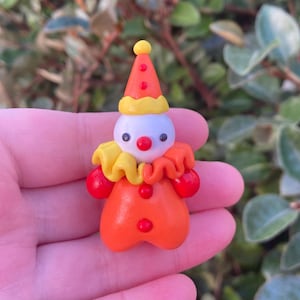 May include: A miniature clown figurine with a white head, red nose, and black eyes. The clown wears an orange outfit with red pom-poms, a yellow ruffled collar, and a red and yellow cone hat. The figurine is handmade.