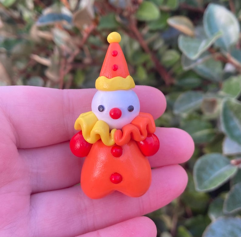 May include: A miniature clown figurine with a white head, red nose, and black eyes. The clown wears an orange outfit with red pom-poms, a yellow ruffled collar, and a red and yellow cone hat. The figurine is handmade.