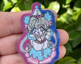 May include: A holographic sticker featuring a cartoon character in a clown-like outfit. The character has gray hair, a sad expression, and is wearing a blue, pink, and white outfit. The sticker has a blue and pink border with star accents.