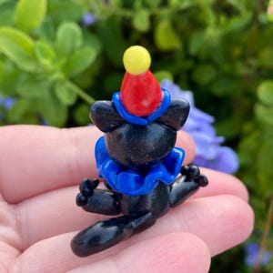 May include: A small, black figurine of a bear wearing a clown outfit. The bear has a red and yellow hat, a blue ruffled collar, and outstretched arms. The figurine is set against a blurred green and purple background.