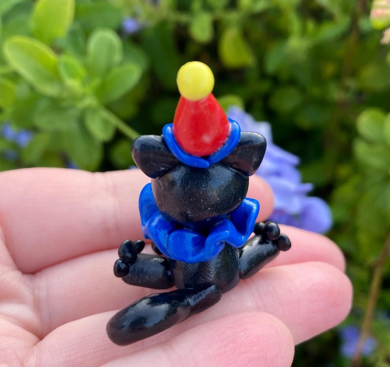 May include: A small, black figurine of a bear wearing a clown outfit. The bear has a red and yellow hat, a blue ruffled collar, and outstretched arms. The figurine is set against a blurred green and purple background.