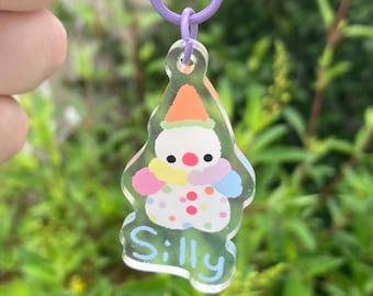 Peut inclure: Un porte-clés en acrylique transparent avec un motif de clown de dessin animé. Le clown a un visage blanc, des yeux noirs et un nez rouge, portant un chapeau coloré. Le mot "Silly" est écrit en bleu clair. Le porte-clés a un anneau violet.