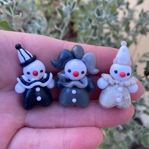 May include: Three small, hand-crafted clay figurines of clowns. Each clown is wearing a different color outfit: black and white, gray and white, and white with a shimmery finish. Each clown has a white face with red lips and a red nose.