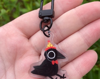 Peut inclure: Un porte-clés en forme de corbeau noir avec un fermoir noir. L'illustration du corbeau porte un nœud papillon rouge et un chapeau de fête jaune à pois rouges. Le porte-clés est en acrylique transparent.