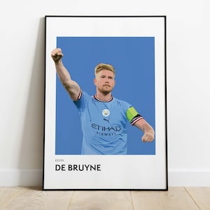Puede incluir: Un estampado enmarcado que presenta una ilustración estilizada de Kevin De Bruyne con una camiseta de fútbol azul claro con el logotipo de Etihad Airways. El jugador está representado con el brazo levantado sobre un fondo azul. El nombre "DE BRUYNE" está impreso debajo.