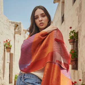 Op de afbeelding: Een kleurrijke sjaal met een geometrisch patroon in tinten oranje, rood en paars. De sjaal is over de schouders gedrapeerd. De sjaal heeft een gefranjerde rand.