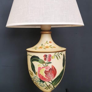 May include: A cream-colored table lamp with a white linen lampshade. The lamp base is decorated with hand-painted floral designs in shades of pink, red, yellow, and green.