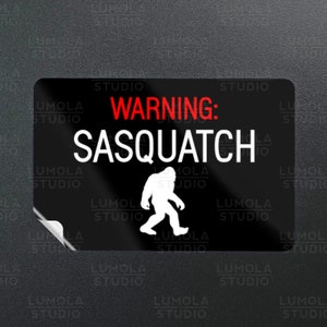 Puede incluir: Pegatina de advertencia en blanco y negro con el texto "WARNING: SASQUATCH" en letras rojas. Una silueta blanca de Bigfoot camina hacia la derecha.