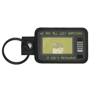 以下が含まれることがあります： 黒いキーホルダー。長方形のパッチには、電子レンジの中にいる漫画のハムスターが描かれています。パッチには「WE ARE ALL JUST HAMSTERS」と「IN GOD'S MICROWAVE」の文字が書かれています。