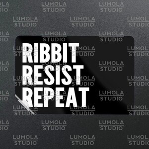 May include: Black sticker with the words "RIBBIT RESIST REPEAT" in bold white letters. The sticker has rounded corners and is surrounded by the text "LUMOLA STUDIO" in a repeating pattern.