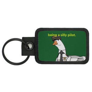 May include: A black and green key chain with a white goose wearing a pilot's hat and uniform. The text "being a silly pilot." is printed on the key chain.