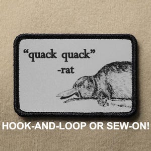 May include: A black and white embroidered patch featuring a platypus and the text "quack quack" -rat. The patch is on a grey background and has a black border. The text "HOOK-AND-LOOP OR SEW-ON!" is below the patch.