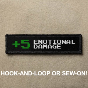 May include: Black and green embroidered patch with the text "+5 EMOTIONAL DAMAGE" in white. The patch is designed to be attached with hook-and-loop or sewn on.