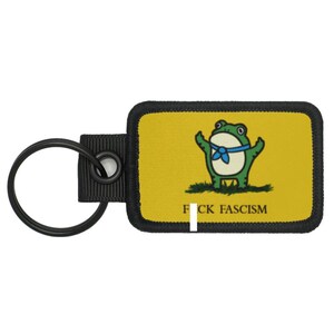 May include: Keyring with a yellow rectangular patch featuring a cartoon frog with a blue neckerchief, arms raised, standing on grass. The text "F*CK FASCISM" is printed below the frog. The keyring has a black metal ring and a black fabric loop.