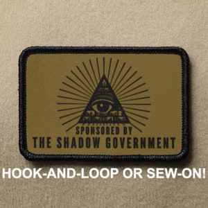May include: A tan and black patch with a black all-seeing eye inside a triangle with rays of light emanating from it. The text "SPONSORED BY THE SHADOW GOVERNMENT" is below the eye. The patch is labeled "HOOK-AND-LOOP OR SEW-ON!"