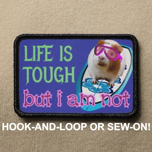 May include: A blue and purple patch featuring a white guinea pig wearing pink sunglasses and riding a surfboard. The text on the patch reads "LIFE IS TOUGH but i am not".