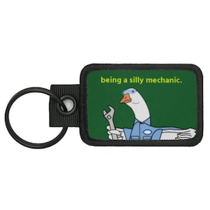 May include: A black and green key chain with a white goose wearing a blue mechanic's uniform and holding a wrench. The text "being a silly mechanic." is printed on the key chain.