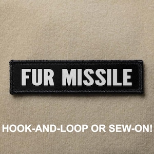 Pode incluir: Patch retangular preto com as palavras "FUR MISSILE" em letras brancas em negrito. O patch é contornado com costura preta. Abaixo do texto está a frase "HOOK-AND-LOOP OR SEW-ON!"