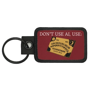 Könnte beinhalten: Ein rechteckiger Schlüsselanhänger mit burgunderrotem Hintergrund und dem Text "DON'T USE AI, USE:". Er zeigt ein Miniatur-Ouija-Brett-Design mit Buchstaben, Zahlen und den Wörtern "YES" und "NO". Ein schwarzer Schlüsselring ist befestigt.