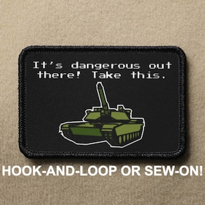 Op de afbeelding: Zwarte en groene geborduurde patch met een cartoon tank en de tekst "It's dangerous out there! Take this." De patch heeft zwarte stiksels rond de randen en staat onderaan "HOOK-AND-LOOP OR SEW-ON!"