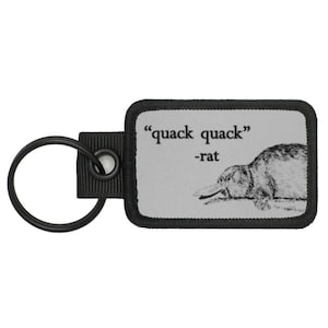 May include: A black key chain with a grey rectangular patch featuring a black and white illustration of a platypus and the text "quack quack" -rat.