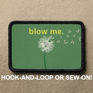 May include: Green fabric patch with black stitching, featuring a white dandelion with seeds blowing away and the text "blow me."