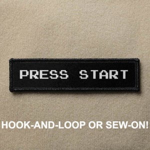 May include: A black rectangular patch with the words "PRESS START" in white pixelated font. The patch is bordered with black stitching and the text "HOOK-AND-LOOP OR SEW-ON!" is below the patch.