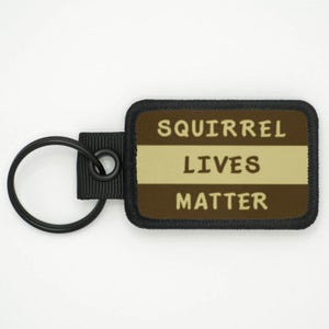 Può includere: Un portachiavi nero e marrone con un cinturino nero e un'etichetta rettangolare marrone che dice "Squirrel Lives Matter".