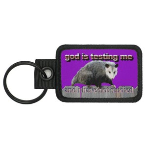 May include: A keychain with a rectangular patch showing a possum illustration on a purple background. The text "god is testing me and I have not studied" is above and below the possum image. The keychain has a black ring.