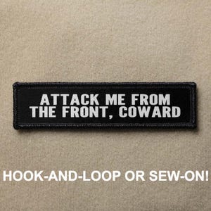 May include: Black and white patch with the text "ATTACK ME FROM THE FRONT, COWARD". The patch is designed to be attached with hook-and-loop or sewn on.