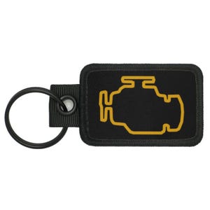 Może przedstawiać: Czarny brelok z żółtym symbolem kontrolki silnika. Brelok jest wykonany z tkaniny i ma metalowy pierścień.