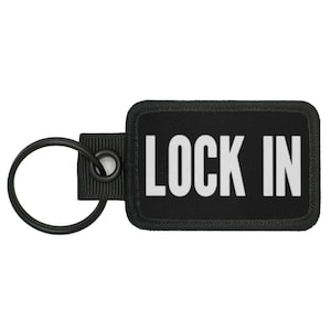 May include: Black rectangular keychain with the words "LOCK IN" in white block letters. The keychain has a black metal ring and a black fabric loop for attachment.