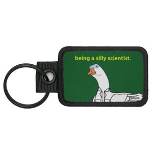 May include: Keyring with a rectangular patch featuring a cartoon goose wearing a lab coat and glasses. The patch has a green background with the text "being a silly scientist." The keyring has a black metal ring and a black fabric loop.