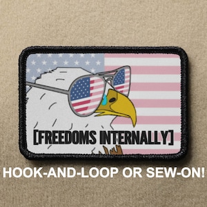Puede incluir: Un parche bordado en blanco y negro con un águila calva de dibujos animados que lleva gafas de sol con el patrón de la bandera estadounidense. El águila mira hacia la derecha y tiene una lágrima que le cae del ojo. El texto "[FREEDOMS INTERNALLY]" está debajo del águila. El parche está sobre un fondo marrón.