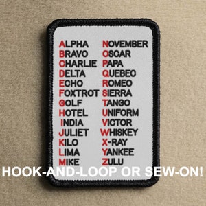 May include: A white patch with black edging features the NATO phonetic alphabet in red and black lettering. The text reads "ALPHA, BRAVO, CHARLIE, DELTA, ECHO, FOXTROT, GOLF, HOTEL, INDIA, JULIET, KILO, LIMA, MIKE, NOVEMBER, OSCAR, PAPA, QUEBEC, ROMEO, SIERRA, TANGO, UNIFORM, VICTOR, WHISKEY, X-RAY, YANKEE, ZULU". The bottom of the patch reads "HOOK-AND-LOOP OR SEW-ON!"