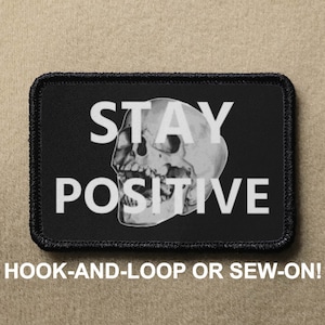 Pode incluir: Patch preto com texto branco que diz "STAY POSITIVE" e um gráfico de caveira. O patch é para fixação de gancho e laço ou para costurar.