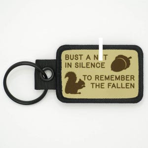 Può includere: Un portachiavi nero e marrone con un design di scoiattolo marrone e ghianda. Il testo sul portachiavi recita "Bust a nut in silence to remember the fallen".