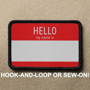 Puede incluir: Parche de etiqueta de nombre rojo y blanco con costuras negras. La etiqueta dice "HELLO my name is" con un espacio en blanco para escribir un nombre. El parche se puede sujetar con velcro o coser.
