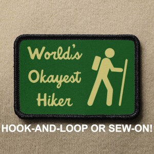 May include: Green and black embroidered patch with a hiker silhouette and the text "World's Okayest Hiker". The patch is for hook-and-loop or sew-on attachment.